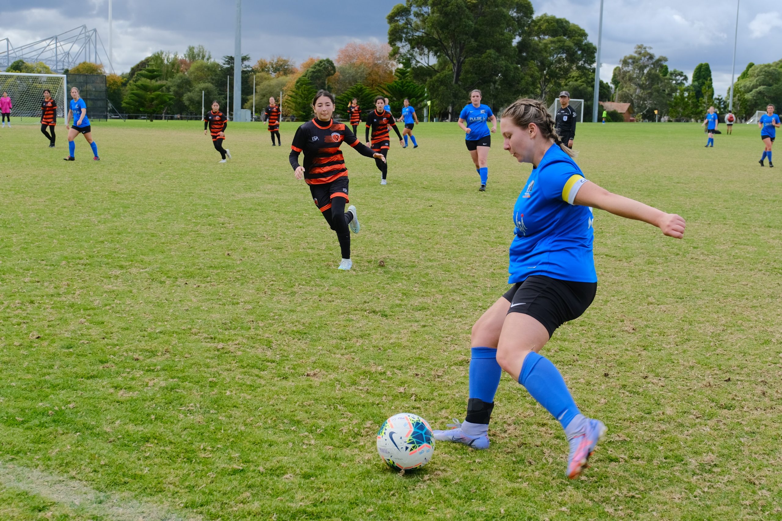 Daisy in the foreground, on the soccer pitch, kicking a ball during a game. She is wearing a yellow and white captain's armband.