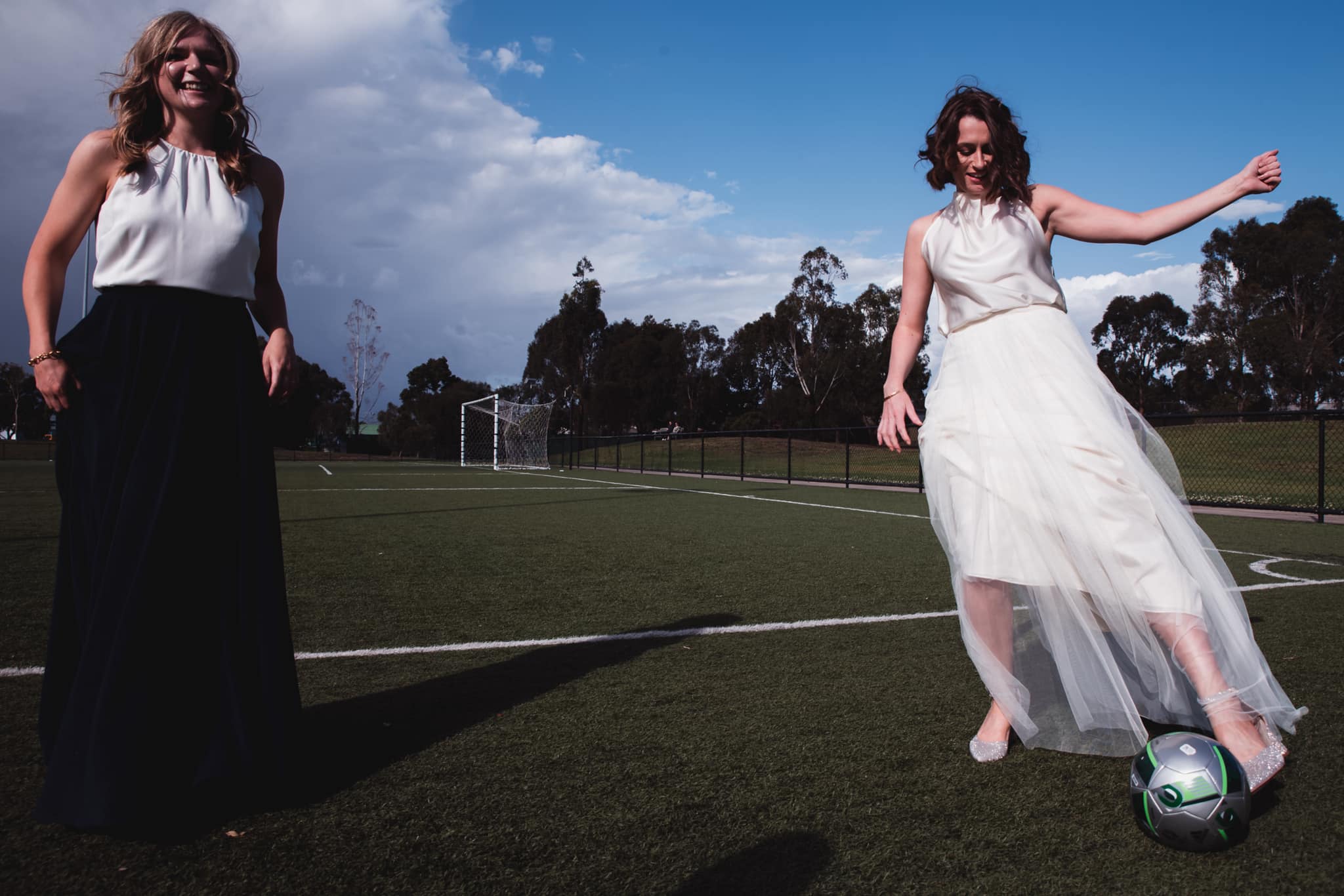 Audrey is in her wedding dress on the right, kicking a soccer ball. Keryn is to the left, smiling. They are at Clifton Park.