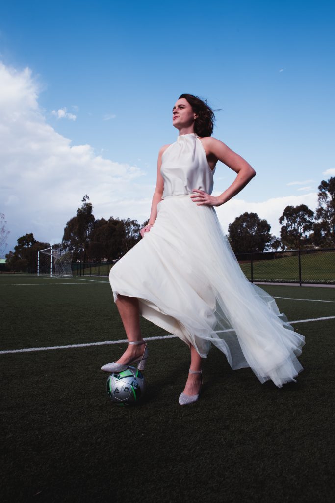 Audrey is standing with one hand on her hip, in her wedding dress, with one heeled foot on a soccer ball at Clifton Park synthetic.