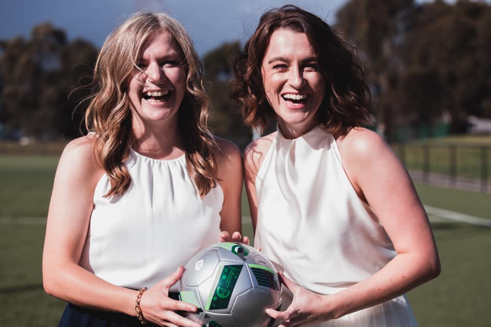 Audrey and Keryn in their wedding dresses, laughing and holding one soccer ball together. 