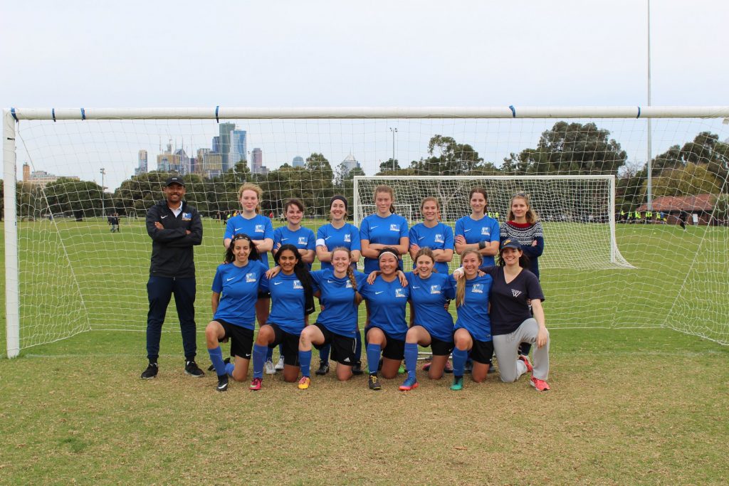 A group photo of the 2018 women's state 3 team, in royal blue home kits, in front of a goal at Princes Park.  Adam, the coach is in black, standing to the left of the group.  Carrot is in the back row of two, on the far left.  