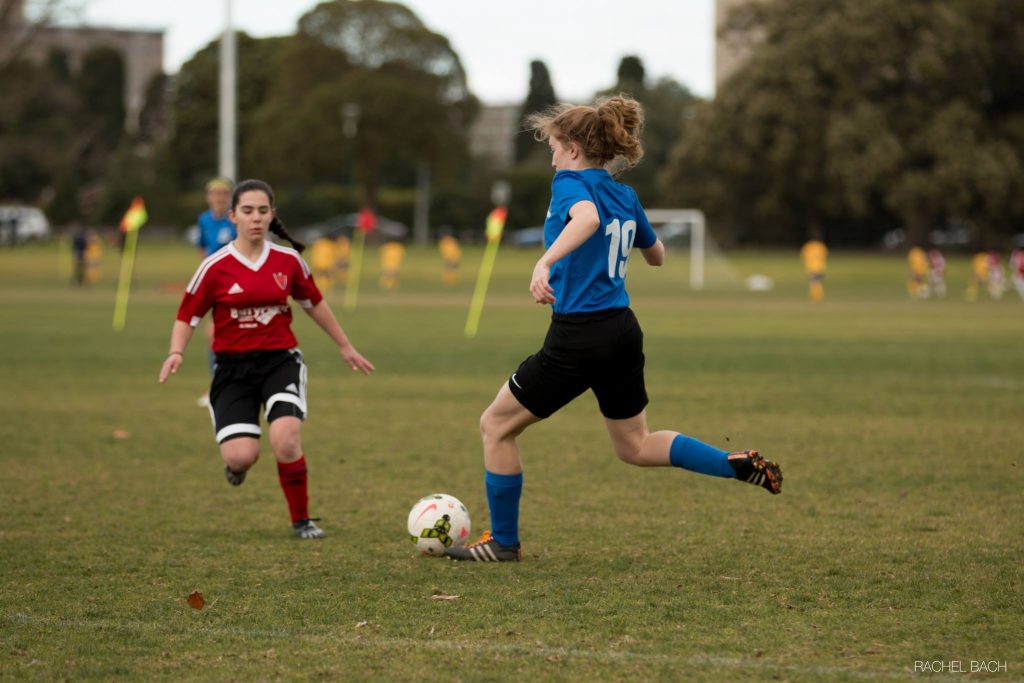 Carrot kicking a ball at Princes Park.  There is an opposition player in red, facing her, and Carrot is wearing MUSC's home kit of royal blue, facing away from the camera.