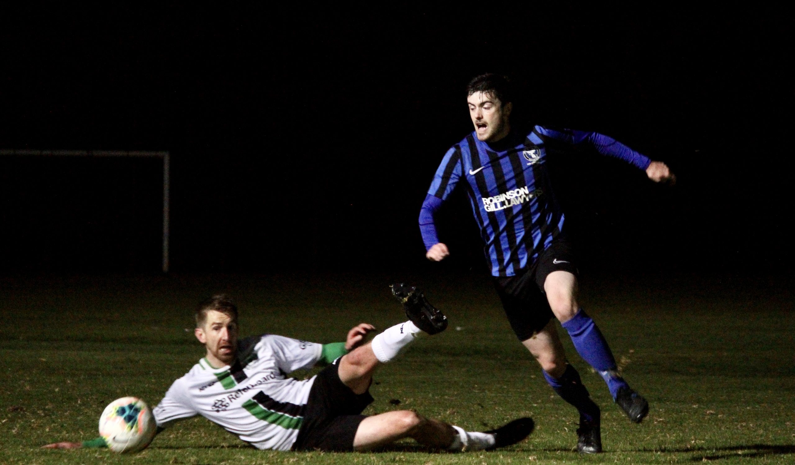 Mitch in a blue and black striped MUSC is on the right side of the photo, running towards a ball in the left bottom corner. An opposition player in a white top is laid on the floor, legs splayed, helpless.